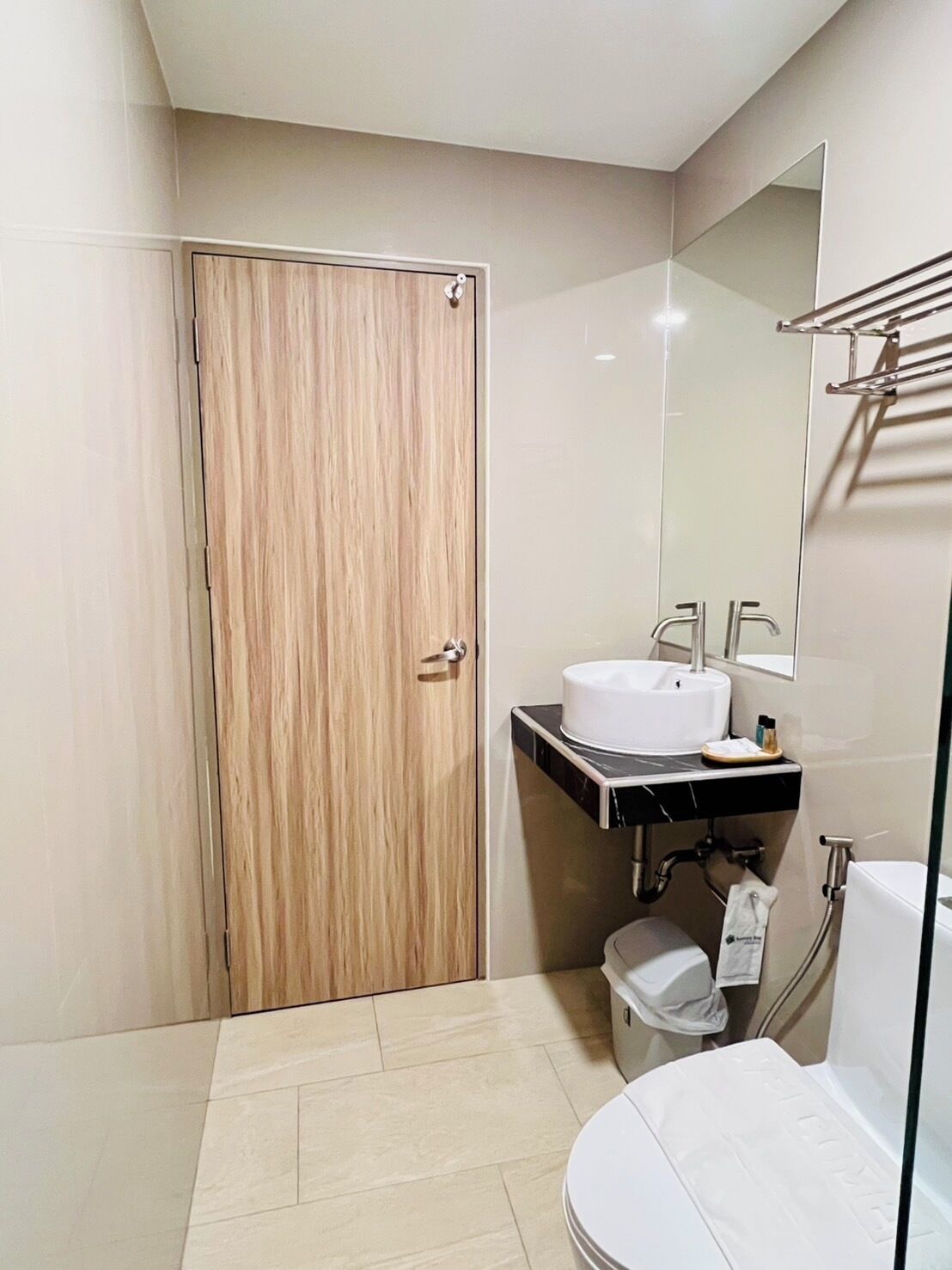 Un baño moderno con puerta de madera, lavamanos blanco, espejo y retrete.