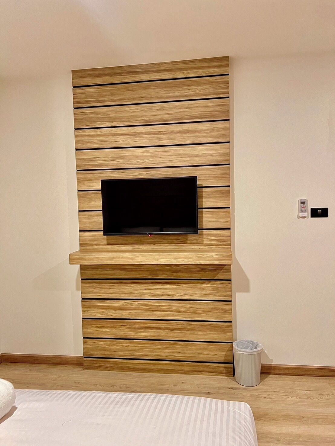 Une chambre moderne avec un téléviseur encastré dans un mur recouvert de panneaux en bois, une poubelle et un lit avec une couette blanche.