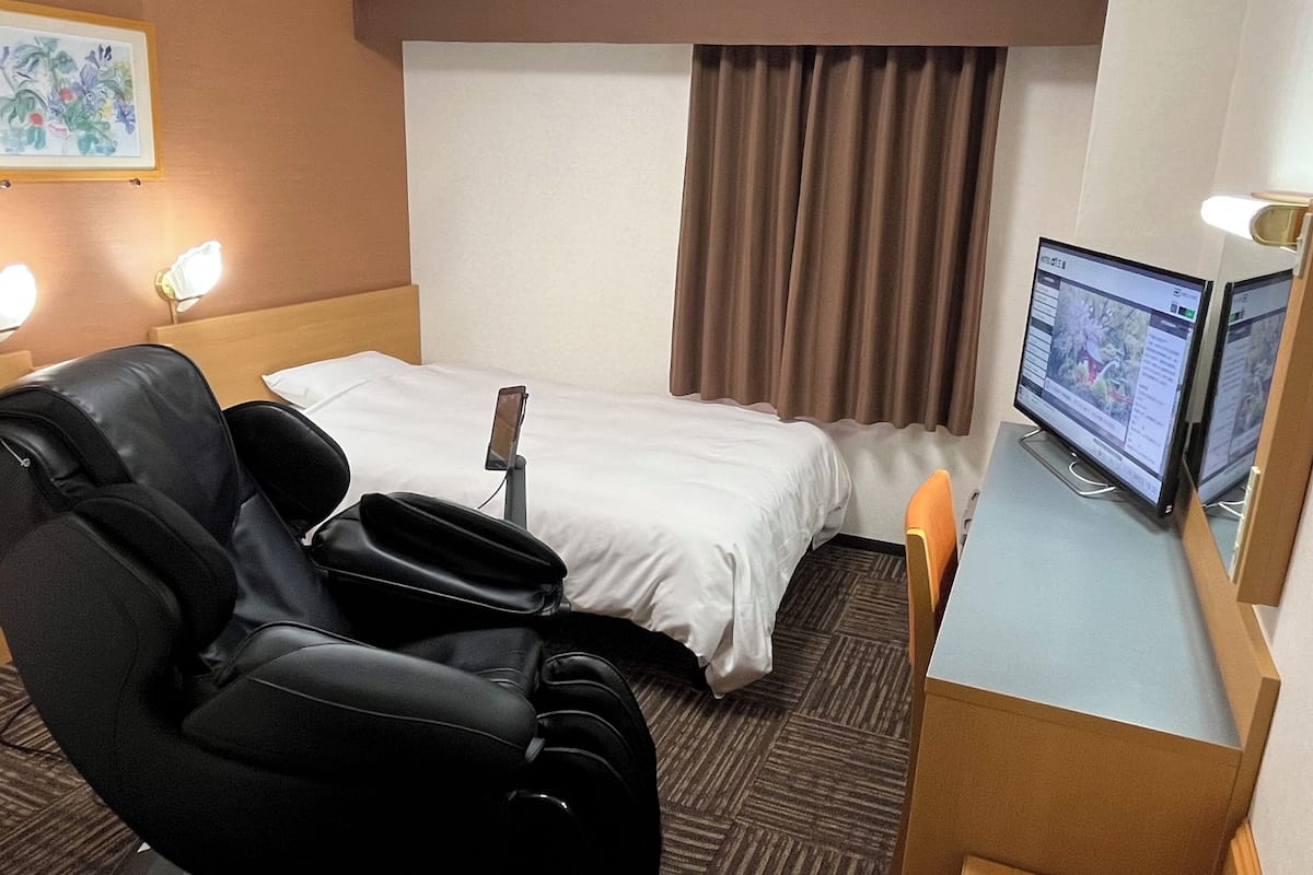 A hotel room with a bed, a desk with a computer, a chair, and a massage chair.