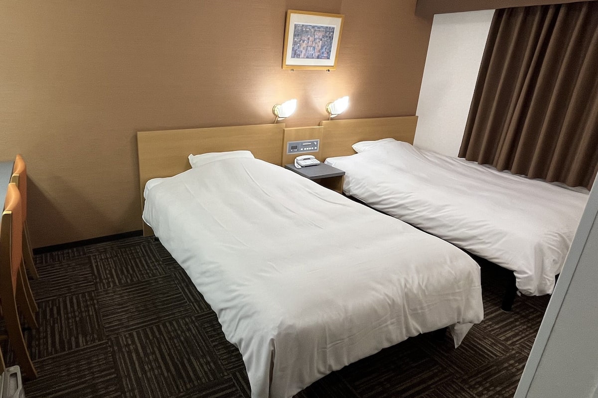 A hotel room with two single beds, a desk, a chair, a telephone, and a framed picture on the wall.