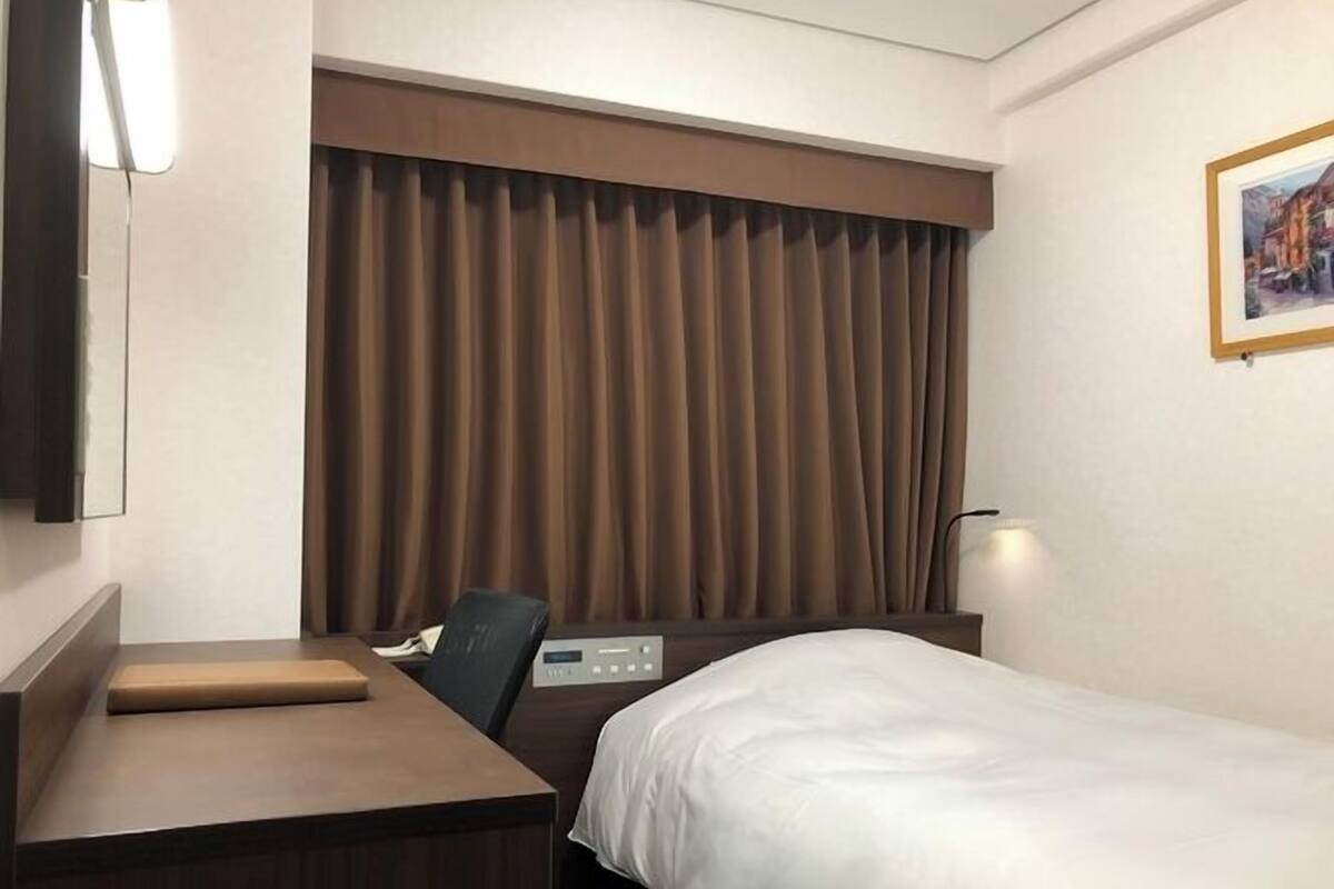 A hotel room with a bed, a desk, a chair, a control panel, and a large window with curtains.