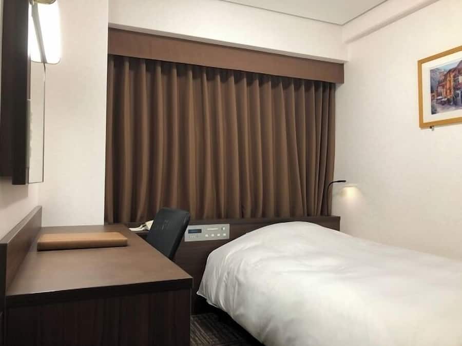 Ein Hotelzimmer mit einem Bett, einem Schreibtisch, einem Stuhl, einer Bedienkonsole und einem großen Fenster mit Vorhängen.