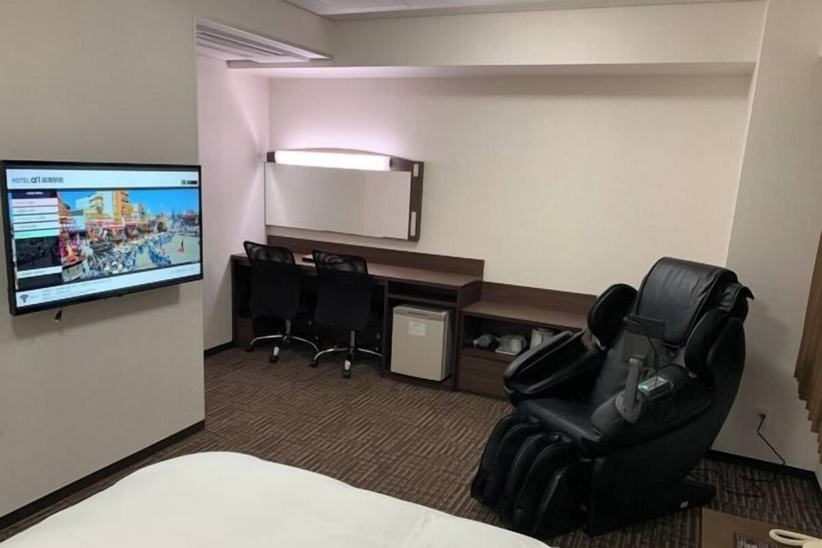 A hotel room with a bed, a desk with two chairs, a massage chair, a television, and a small refrigerator.