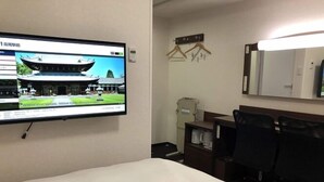 Habitación de hotel con cama, escritorio, dos sillas, un televisor mostrando un edificio y un perchero.