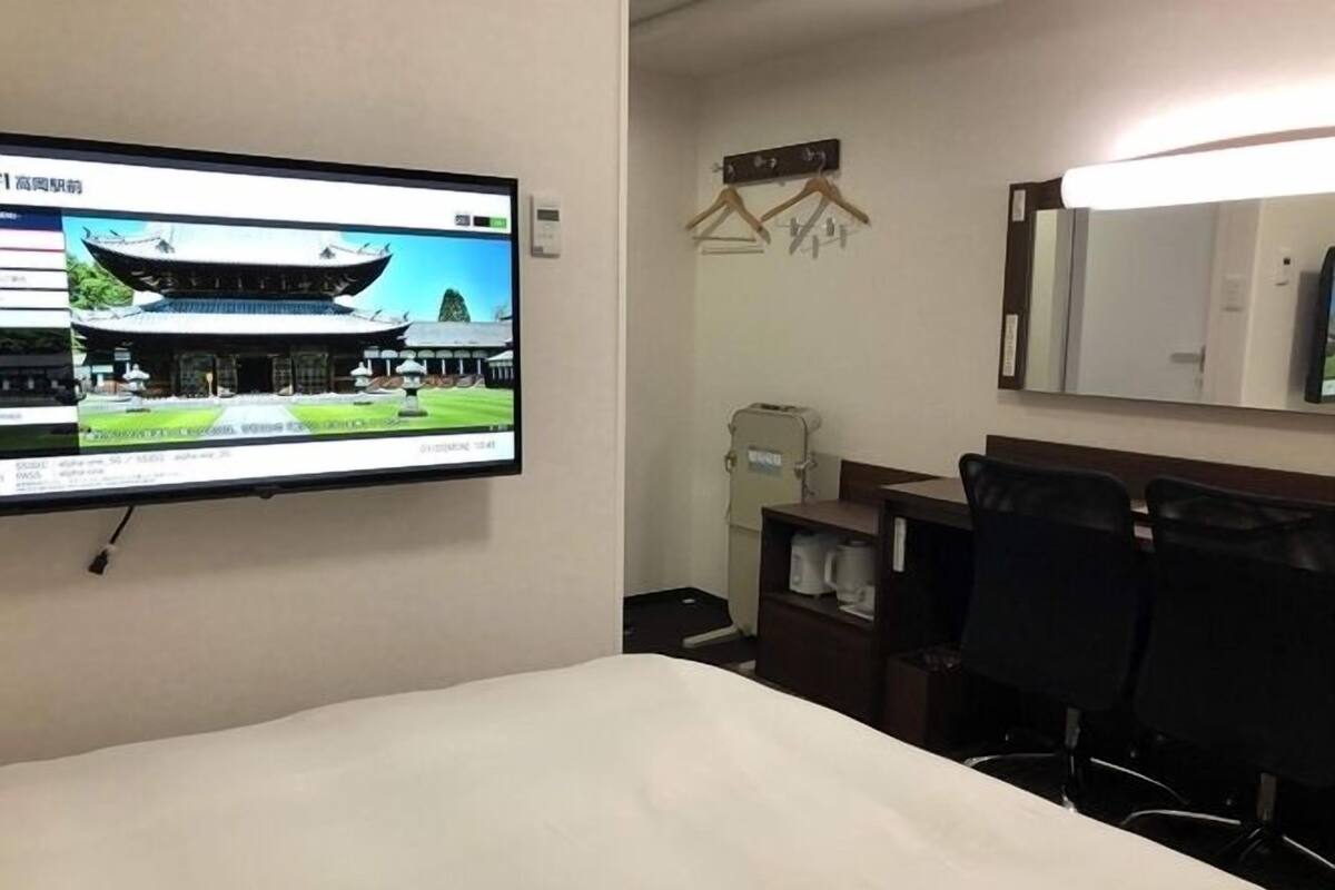 A hotel room with a bed, a desk, two chairs, a television displaying a building, and a coat rack.