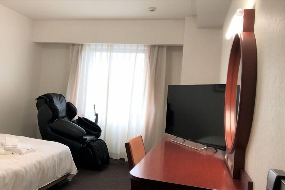 A hotel room with a bed, a desk with a flat-screen TV, a black leather chair, and a large mirror.