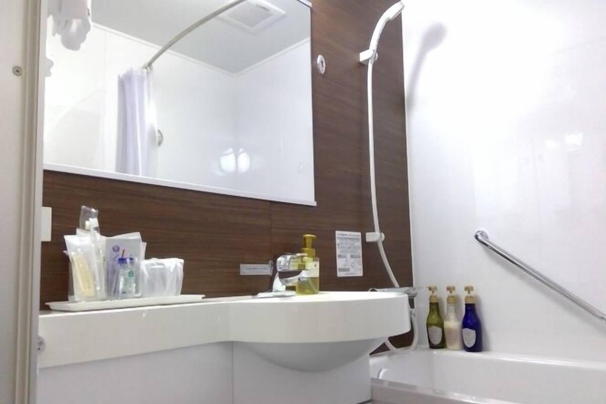 Un baño con un lavabo blanco, espejo y ducha. Hay artículos de aseo en el lavabo y un asidero cerca de la ducha.