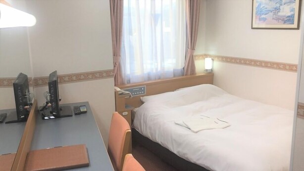 Habitación de hotel con cama, escritorio, computadora, silla y ventana con cortinas.