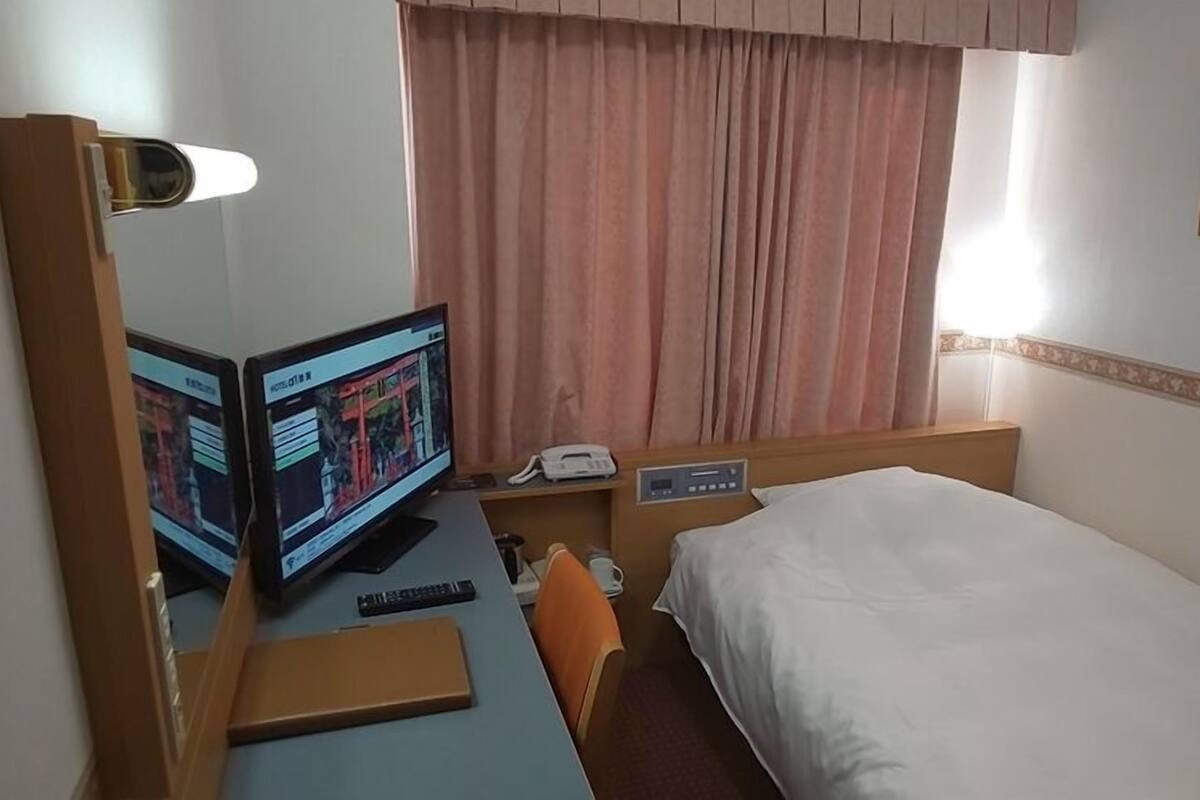 Une chambre d’hôtel avec un lit, un bureau avec un ordinateur, un téléphone et une fenêtre avec des rideaux.