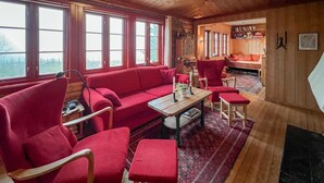 A cozy room with a red sofa, matching armchairs, and a wooden table. Large windows provide natural light, and a bookshelf is visible in the background. - Awesome home in Lillehammer with sauna (Lillehammer)