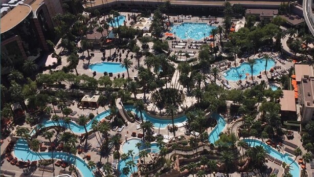 A large outdoor swimming pool area with multiple interconnected pools, surrounded by palm trees and lounge chairs.