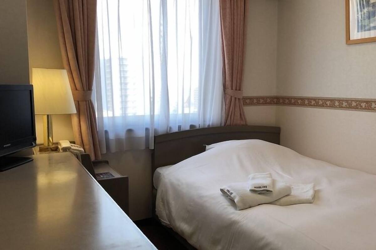 A hotel room with a bed, a desk with a lamp, a television, and a window with curtains.