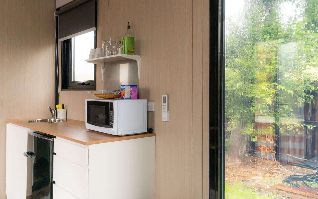 Cocina compacta con microondas, un lavamanos pequeño y ventana con vistas a zona boscosa.