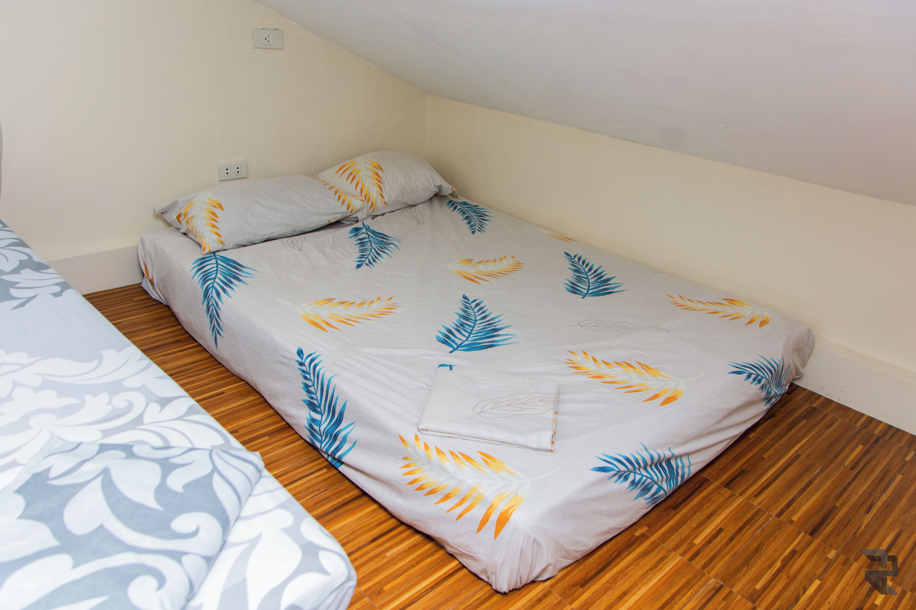 Ein Einzelbett mit Bettdecke im Blattmuster in einem Zimmer mit Holzboden und weißen Wänden.