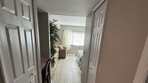 Um corredor estreito com portas brancas de ambos os lados, conduzindo a um quarto com cama e janela.