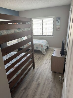 A bedroom with a bunk bed, a bedside table, a window with blinds, and a wall-mounted TV. - Ocean View Condo • Pool • Beachy Getaway (Cocoa Beach)
