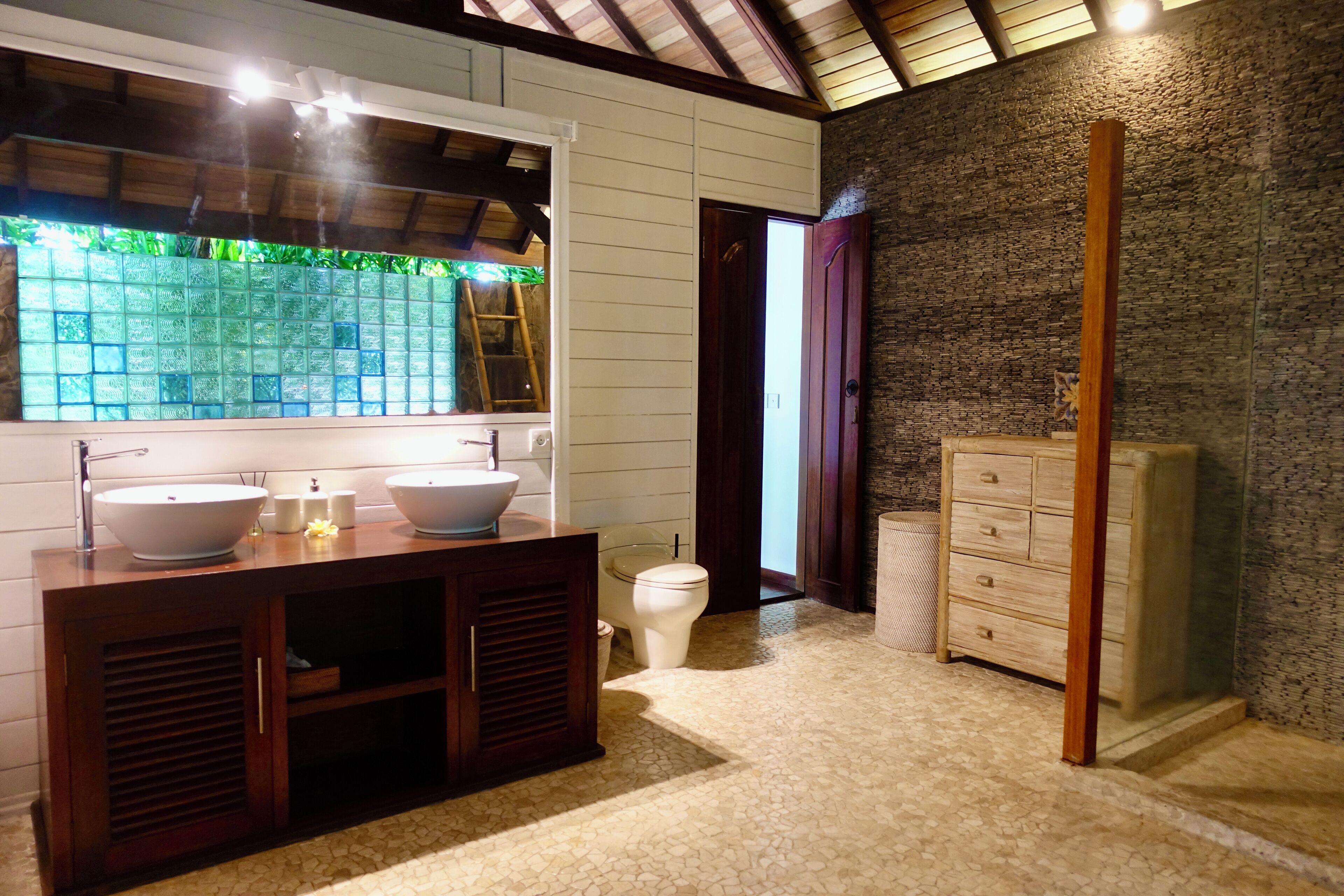 A bathroom with two sinks, a toilet, a wooden cabinet, and a stone wall.