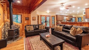 A log cabin interior with a fireplace, leather sofas, a coffee table, and a kitchen area. - Summer Lodge Life! 5bd+4bath+HotTub+Sauna+EV (Ashford)