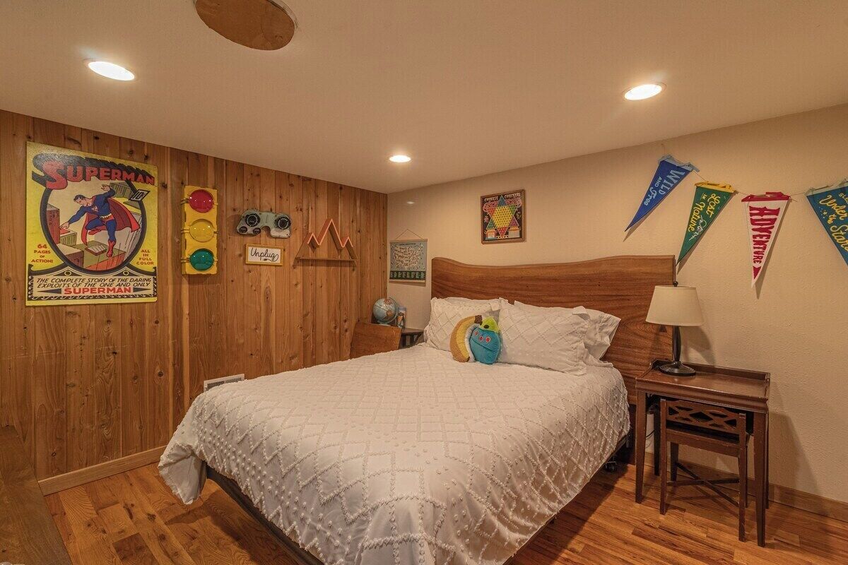 Une chambre à coucher avec une tête de lit en bois, un lit recouvert de linge de lit blanc, une table de chevet avec une lampe, et des décorations murales comprenant une affiche de Superman et des fanions.