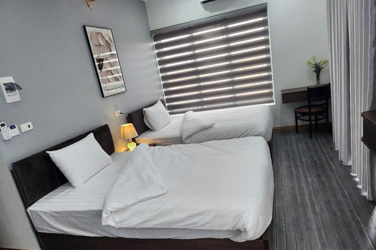A double bed with white bedding, a bedside table with a lamp, a framed picture on the wall, and a window with blinds.