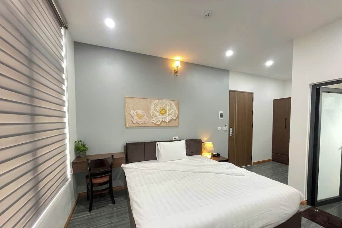 A modern hotel room with a large bed, a desk, and a chair. There is a wall-mounted artwork above the bed and a door to the right.