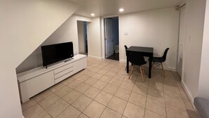 Een kleine, goed verlichte kamer met een televisie op een wit kastje, een eettafel met stoelen en een deur naar een andere ruimte.