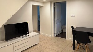 Een moderne, goed verlichte kamer met een flatscreen televisie op een witte tv-meubel, een eettafel met een zwarte stoel, en een deur naar een andere kamer.