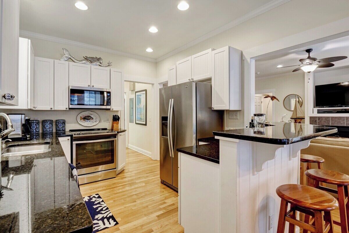 A modern kitchen with white cabinets, stainless steel appliances, a wooden floor, and a bar area with stools.
