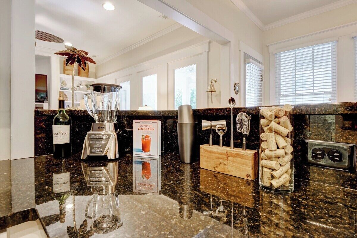 A kitchen with a granite countertop, a blender, a bottle of wine, a wooden cutting board, and a glass jar filled with corks.