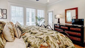 A bedroom with a bed, a ceiling fan, a dresser, a TV, and large windows.