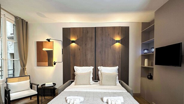Een hotelkamer met een groot bed, twee nachtkastjes, een stoel, een aan de muur bevestigde televisie en een raam met gordijnen.