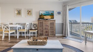 Un salon spacieux doté d’un grand téléviseur, de meubles blancs et offrant une vue sur la plage grâce à une porte-fenêtre coulissante.
