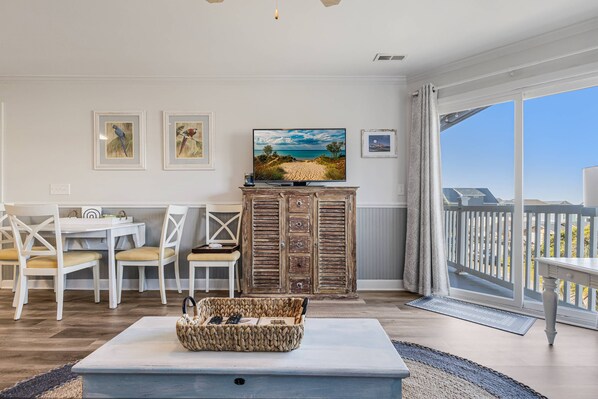 A spacious living room with a large TV, white furniture, and a view of the beach through the sliding glass door.