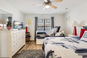 Une chambre à coucher comprenant un lit, une commode, un ventilateur de plafond, une fenêtre avec des rideaux et une vue sur des immeubles.