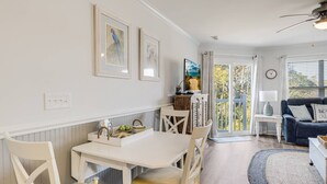 Un coin repas avec une table et des chaises blanches, une petite table d’appoint avec une lampe, et une vue sur l’extérieur grâce à une porte vitrée.