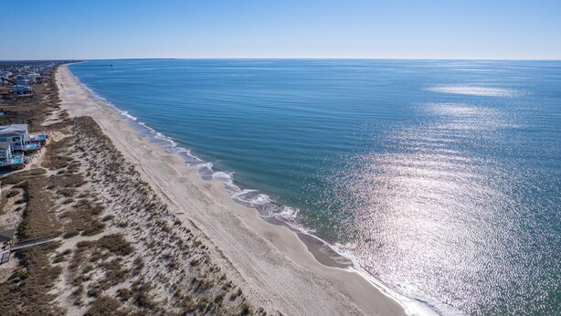Une vue aérienne d’une plage, avec un ciel dégagé, une côte sablonneuse et quelques bâtiments le long de la côte.