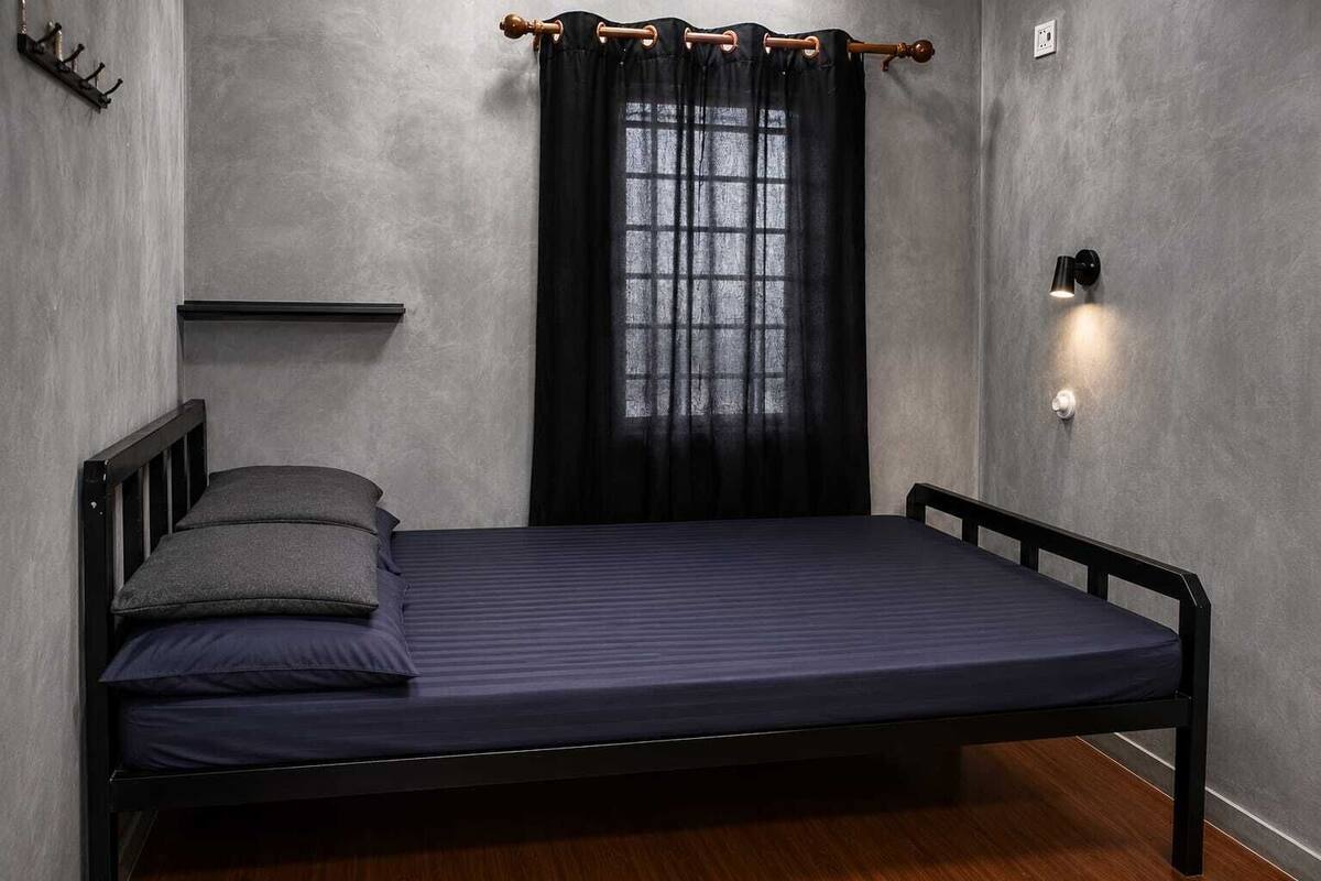 A single bed with a headboard, a bedside table, a wall-mounted light, and a window with a curtain.