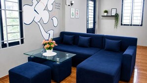A modern living room with a blue sectional sofa, a glass coffee table with a flower centerpiece, and a wall with abstract art and motivational quotes. - The Drop Inn (Siem Reap)