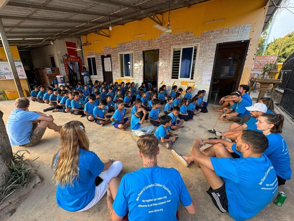 A group of children and adults seated in an outdoor area with a yellow wall and brickwork, under a metal roof structure.