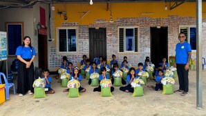 A group of children seated on stools with bags, in an outdoor setting with a yellow wall and brick pattern. - The Drop Inn (Siem Reap)
