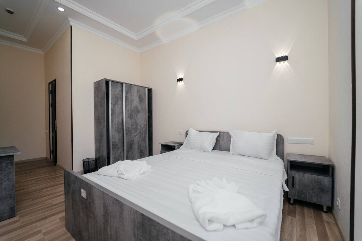 Ein modernes Hotelzimmer mit einem großen Bett, Holzboden und einem Kleiderschrank. Zwei Nachttische und eine Wandleuchte sind vorhanden.