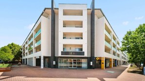 A multi-story building with a central entrance, balconies, and a brick-paved ground. - Lakeside 1-Bed with A/C, Parking & Balcony (Greenway)