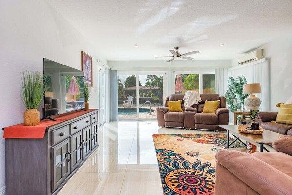 A spacious living room with a sofa, a coffee table, and a TV. There is a large window with a view of a pool and outdoor furniture. - Juanita's Waterfront Villa (Fort Lauderdale)