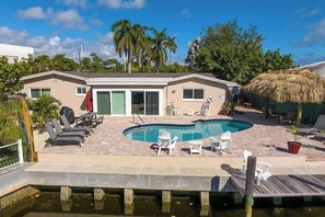 A pool area with lounge chairs, a thatched roof structure, and a house with a green door. - Juanita's Waterfront Villa (Fort Lauderdale)