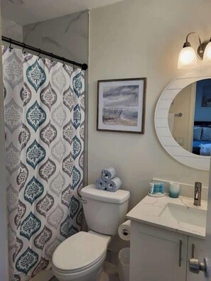 A bathroom with a patterned shower curtain, a framed picture on the wall, a toilet with toilet paper, and a sink with toiletries.