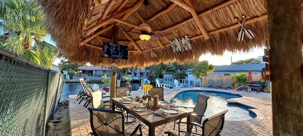 Un espacio de comedor al aire libre con techo de paja, piscina y vistas de los edificios circundantes.