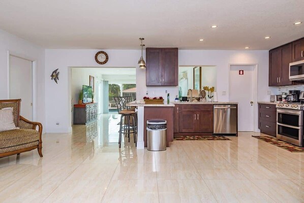 A spacious living room with a sofa, a dining area with chairs, a kitchen with modern appliances, and a large window.