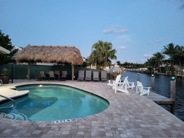 A swimming pool with a thatched roof structure, lounge chairs, and a dock extending into a body of water.