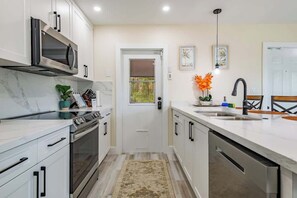 A modern kitchen with white cabinets, a built-in microwave, a stove, and a dishwasher. There is a door with a window, a sink, and a vase with flowers on the counter.