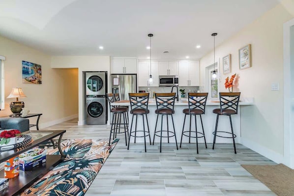 A modern kitchen with white cabinets, a central island with bar stools, a washing machine, and a microwave.